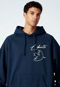 Sweat à capuche bleu marine en mélange de coton, présentant une colombe blanche brodée et le texte « Liberté ». Comprend une poche avant et une capuche avec cordon de serrage.