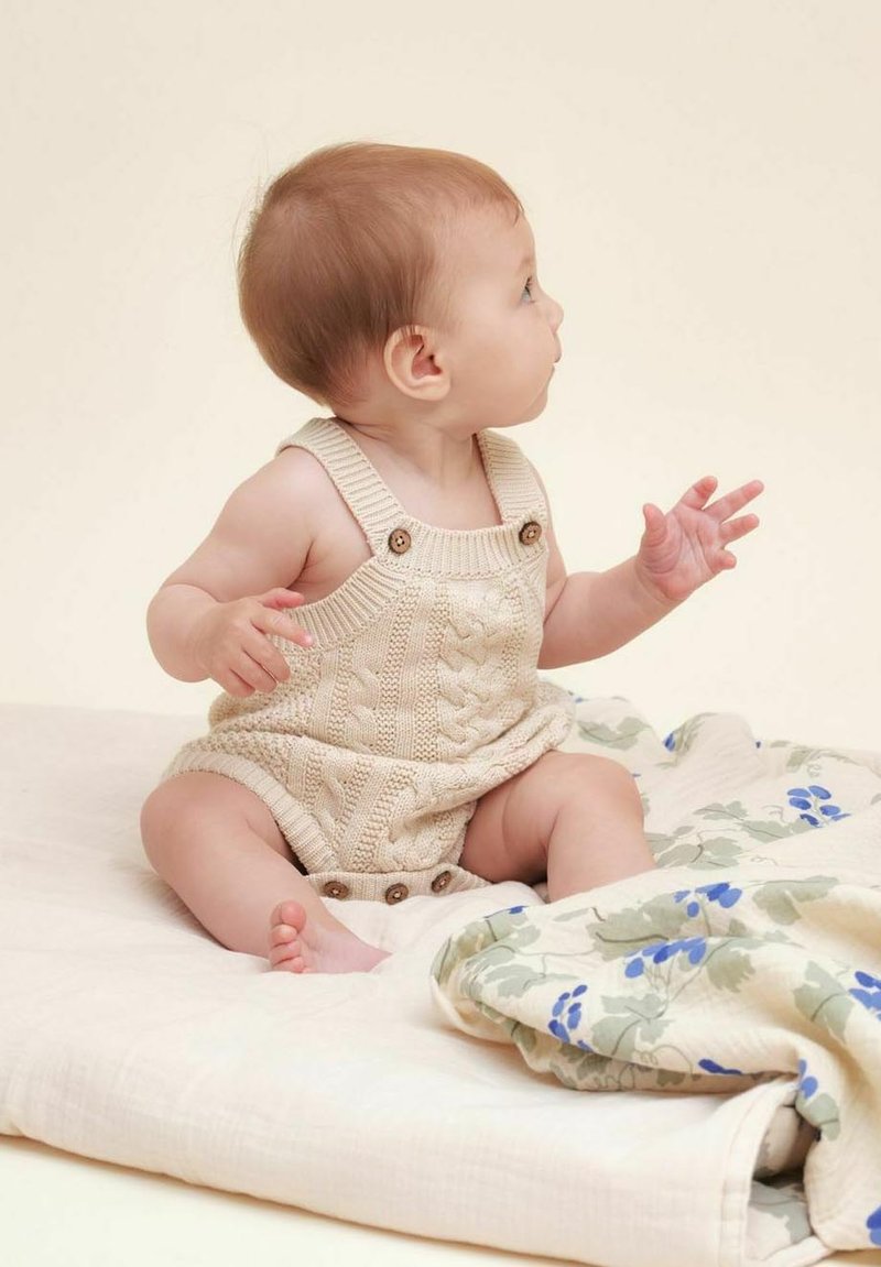 Beige strikket romper med kabelmønster og knapdetaljer, båret af en baby, der sidder på et blødt, mønstret tæppe mod en neutral baggrund.