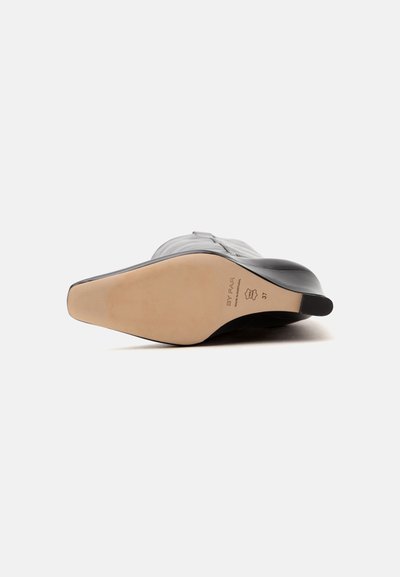 Botte en cuir noir à bout pointu, couchée sur le côté montrant une semelle lisse couleur caramel avec le nom de la marque, une marque en cuir et la pointure 37.