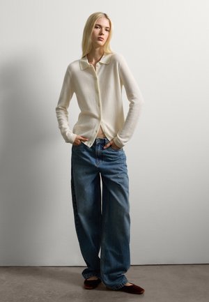 Cardigan button-up bianco sporco con una texture morbida, abbinato a jeans in denim blu larghi. Il modello è in piedi con le mani nelle tasche contro un muro semplice.
