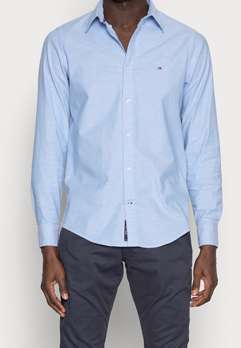 Hombre vestido con una camisa de botones de manga larga azul clara con un pequeño logo en el pecho y pantalones oscuros, de pie contra un fondo liso.