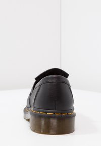 Svart lädersko med rundad form, som har synliga sömmar och en texturerad gummisula med gula accentstickningar.