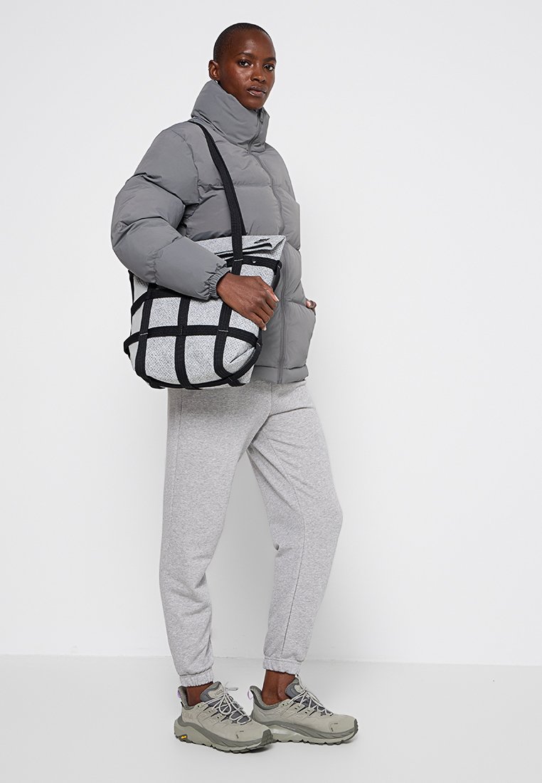 Graue Pufferjacke mit hohem Kragen, kombiniert mit hellgrauen Jogginghosen und grauen Sneakers. Die Tragetasche hat ein schwarzes Gittermuster.