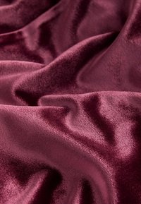Burgundijas samta audums ar gludu, spīdīgu tekstūru, kas parāda mīkstus krokus un gaismas atspīdumus virsmas. 