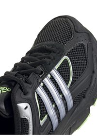 Černá sportovní bota s síťovaným svrškem, výrazným stříbrným designem se třemi pruhy, zelenými akcenty a texturovanou gumovou podešví.