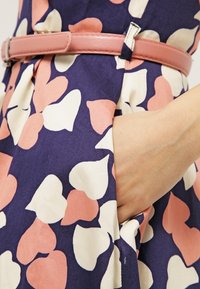 Blumenmusterkleid aus überwiegend navyfarbenem Stoff, mit herzförmigen Motiven in Pfirsich und Weiß. Enthält einen pinken Leder-Gürtel und seitliche Taschen.