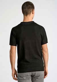 Svart kortärmad polo med krage, med ribbad nederkant och strukturerad stickad tyg. Modellens bakvy visar en ren, figursydd design.