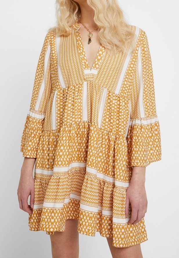 Femme portant une robe ample, mi-genoux, à manches longues avec des motifs géométriques jaune moutarde et blanc, debout devant un fond uni.