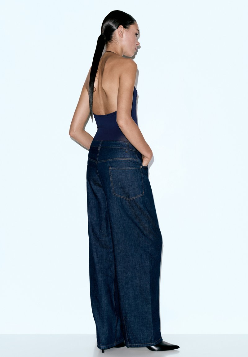 Džíny s vysokým pasem a širokými nohavicemi z tmavého denimu s hladkým povrchem, s zadními kapsami a kontrastním prošitím. Model zobrazen zezadu.