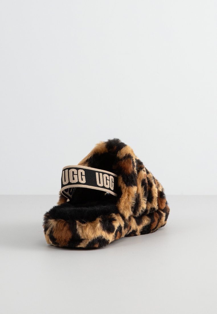 Klasyczne klapki z futra z leopardzim wzorem, z miękką czarną podeszwą i szerokim beżowym paskiem, na którym znajduje się nazwa marki "UGG" w czarnym druku.