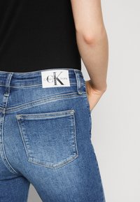 Blue denim jeans with a slight fade, featuring a white Calvin Klein label at the waistband and single back pocket design. Textured fabric.