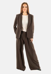 Blazer marrón a medida sobre una blusa blanca con estampado, combinado con unos pantalones marrones de pierna ancha. El conjunto presenta líneas nítidas y un mínimo de herrajes.