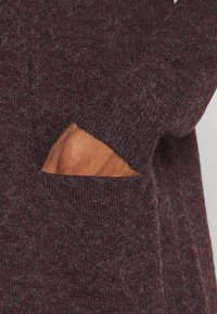 Hand delvis nedstoppad i fickan på en vinröd, fluffig, stickad tröja.