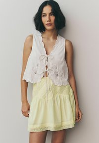 Crop top blanc brodé avec un décolleté à volants, associé à une mini jupe jaune texturée avec des accents en dentelle et un cordon de serrage.