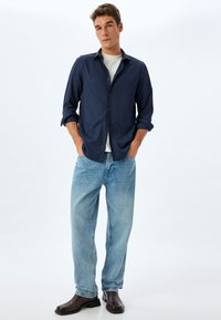 Camisa azul marino de botones, con mangas enrolladas; camiseta blanca de cuello redondo debajo. Jeans de color azul claro, botas marrones hasta el tobillo, fondo neutro.