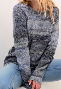 Stickad tröja i nyanser av blått och grått. Strukturerat mönster med långa ärmar och ribbade muddar. Bärs över ljusblå jeans.