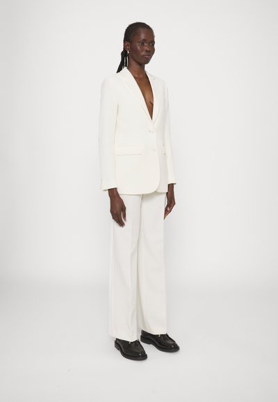 Completo bianco con blazer a profondo scollo a V e pantaloni a gamba larga. Tessuto testurizzato, tasche frontali, abbinato a scarpe nere. Sfondo bianco uniforme.