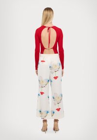 Haut long à manches ribblées rouge avec un dos ouvert et un détail à nouer, associé à un pantalon ample blanc orné d'imprimés floraux bleus et rouges.