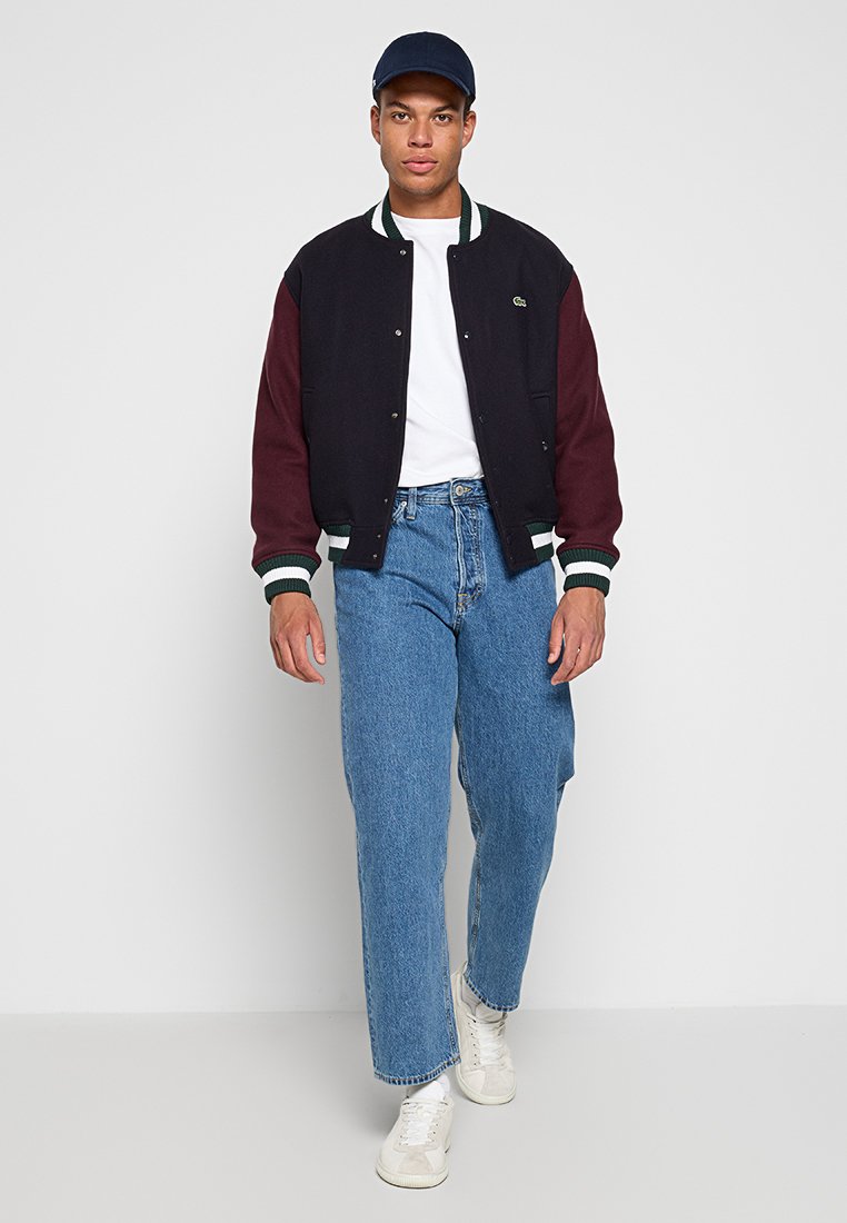 Veste varsity noire et bordeaux avec des accents blancs sur un t-shirt blanc, associée à un jean bleu clair et des baskets blanches.