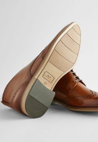 Chaussures brogue en cuir brun avec des détails perforés, dotées d'une semelle beige clair et d'un capuchon de talon en caoutchouc gris. Design simple avec laçage.