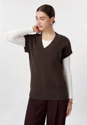 Nő sötétbarna V-nyakú ujjatlan pulóverben, fehér hosszú ujjú felsővel, hozzáillő sötétbarna nadrággal és arany fülbevalóval.