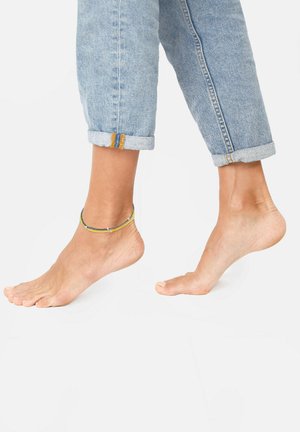 Bracciale alla caviglia con perline verdi, gialle e blu, indossato su un piede nudo. Jeans in denim azzurri chiari arrotolati in modo informale.