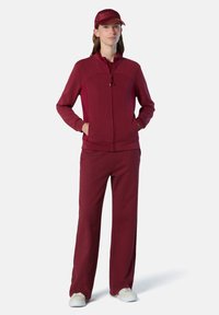 Bordeauxfarbener Sportanzug mit Reißverschlussjacke und weit geschnittenen Hosen. Glatter Stoff, figurbetontes Design mit Stehkragen und Taschen. Weiße Sneakers.