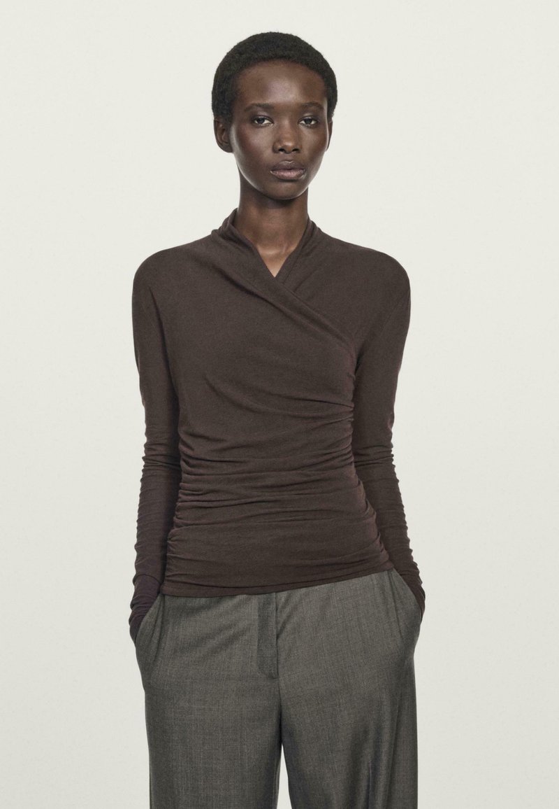 Woman with short hair wearing a fitted dark brown ruched long-sleeve top and loose gray trousers with hands in pockets.