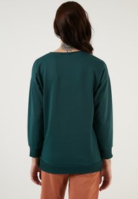 Felpa color teal con una vestibilità comoda, spalle basse e polsini e orlo a costine. Presenta un ampio scollo e una texture di tessuto liscia e morbida.