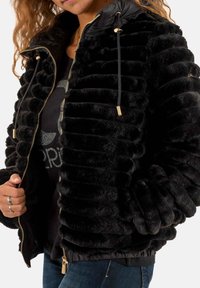 Giacca a vento nera con texture morbida e rigata, chiusura con zip, cappuccio removibile con cordini e dettagli in metallo dorato.