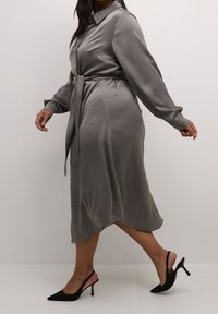 Robe gris à manches longues avec col, taille nouée et texture douce. Associée à des talons slingback noirs. Coupe slim et longueur au genou.