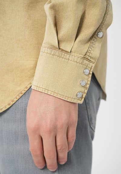 Poignet de chemise beige avec trois boutons-pression métalliques, montrant une texture de tissu lisse et une bordure nette et ajustée. Jean gris partiellement visible.