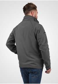 Grijze softshell jas met een hoge kraag, ritszakken en een subtiele logo-accent op de mouw. Beschikt over een gladde textuur en een aangesloten ontwerp.