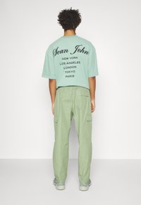 Mintgrön oversized t-shirt med svart text som listar städer på baksidan; kombinerad med ljusgröna cargobyxor med sidofickor.