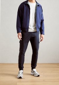 Marineblauwe jas met rits, lichtgrijze T-shirt, donkere cargo-broek en witte sportschoenen met zwarte accenten, staand op een houten vloer.