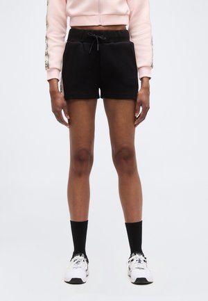 Άτομο που φοράει μαύρο σορτς με κορδόνι, ανοιχτό ροζ ζακέτα με φερμουάρ και μανίκια με σχέδια, μαύρες κάλτσες και λευκά αθλητικά παπούτσια.