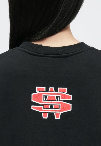 Melna džemperis ar saplaisātu sarkano un baltu grafisko logotipu aizmugurē. Audums izskatās mīksts, ar klasisku piegriezumu un ribotu apkakli.