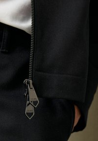 Svart jacka med en jämn yta; har en metallblixtlås med dubbla dragkedjor, graverad logotyp och en slim fit-design.