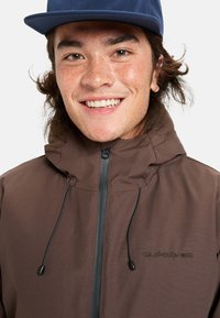 Ung man med brunprickig hud som bär en mörkbrun jacka med huva och svart dragkedja samt en blå keps, leende.