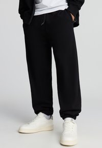 Pantaloni tuta neri con elastico in vita e alle caviglie, realizzati in tessuto morbido. Abbinati a sneakers bianche con dettagli perforati.