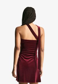 Žena s dlhými vrkočmi oblečená v bezrukávových, jednonohých sametových vínových šatách, otočená chrbtom proti jednoduchému pozadiu.