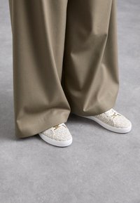 White patterned sneakers with a textured upper and gold hardware, paired with loose, taupe pants, on a gray surface.
