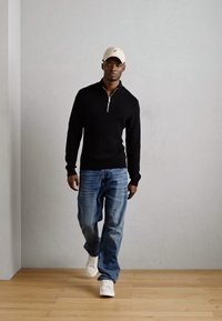 Black zip-neck sweater, blue jeans, and white sneakers. Model wears a beige cap and stands on a light wood floor against a gray wall.