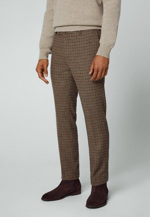 Homme portant un pantalon à carreaux marron, un pull beige et des bottines Chelsea en suède marron foncé, se tenant devant un fond uni.