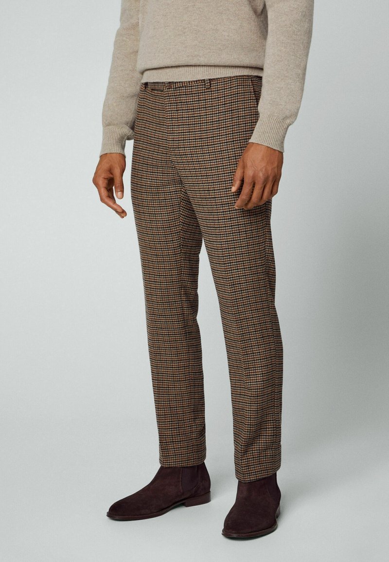 Hnědé kostkované kalhoty s přiléhavým střihem, vyrobené z texturované tkaniny. Doplňuje je tmavě hnědé kotníkové boty a světlejší svetr.