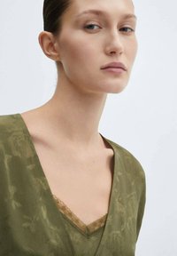 Top vert olive avec un col en V, présentant des motifs floraux et des accents en dentelle au niveau du col, fabriqué en tissu léger.