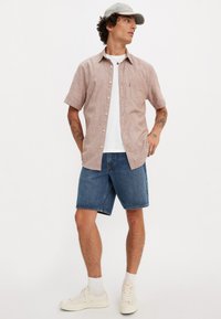 Kurzarm, hellrotes Leinenhemd mit einer Brusttasche, kombiniert mit blauen Jeansshorts und weißen Turnschuhen. Graue Mütze auf dem Kopf.