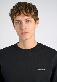 Svart sweatshirt med rund halsringning, med en liten vit "LINDVERGH"-logotyp på bröstet. Slät tygerstruktur, minimalistisk design.