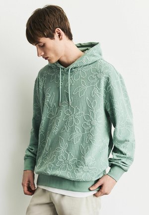 Young man wearing a light green floral textured hoodie with drawstrings, looking down, with beige pants against a plain background.