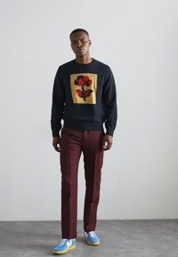 Μαύρος πουλόβερ με λουλουδάτο σχέδιο σε κόκκινο και κίτρινο στο κέντρο, συνδυασμένο με μπορντώ παντελόνι και μπλε αθλητικά παπούτσια με κίτρινες σόλες.